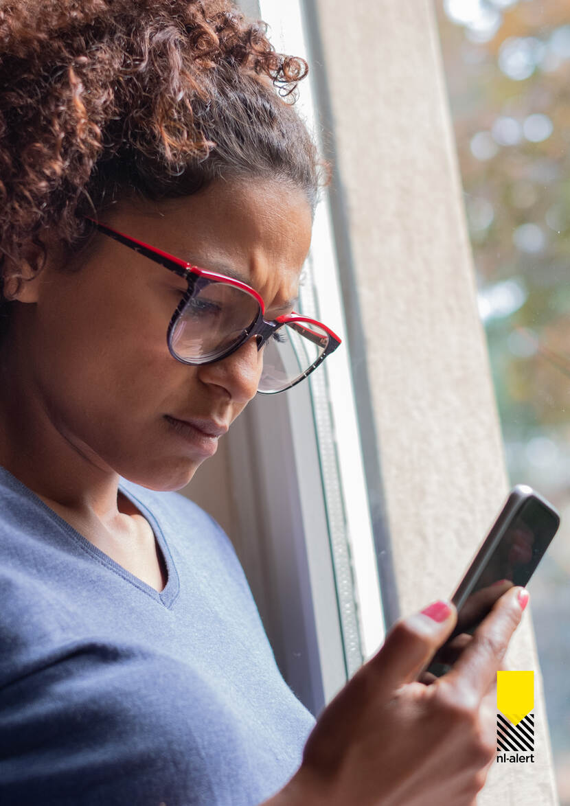 Vrouw met bril op zit staat bij het raam en kijkt op mobiele telefoon in haar hand, het is dag