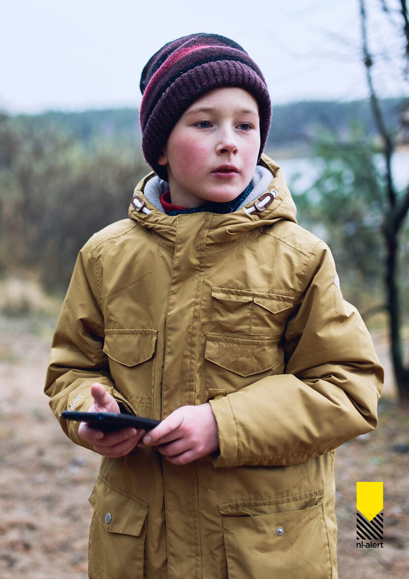 Jongetje in een natuurgebied in de winter met warme jas en muts op staat met mobiele telefoon in zijn hand.
